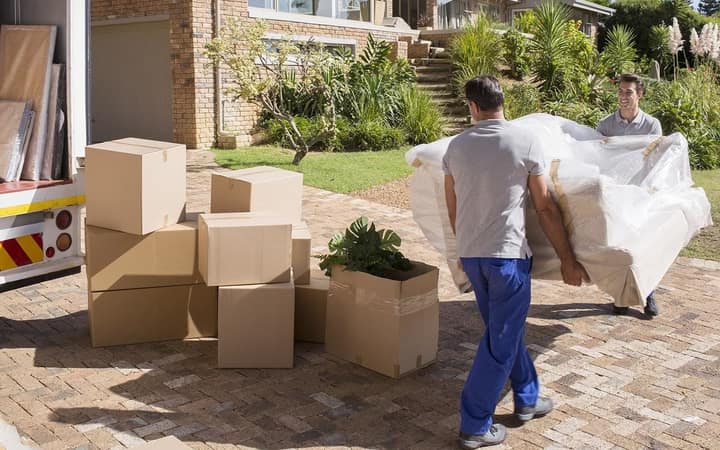 Two movers carrying a wrapped sofa past packed cartons